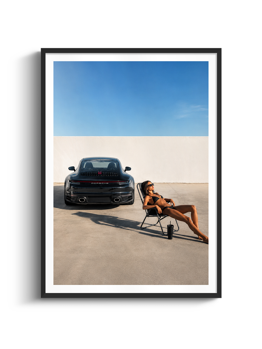 A framed art print featuring a black Porsche 911 sports car parked in a minimalist concrete courtyard under a clear blue sky, with a woman lounging in a chair nearby. The print is presented in a white mat and a sleek black frame against a clean white background.