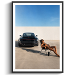 A framed art print featuring a black Porsche 911 sports car parked in a minimalist concrete courtyard under a clear blue sky, with a woman lounging in a chair nearby. The print is presented in a white mat and a sleek black frame against a clean white background.