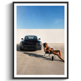 A framed art print featuring a black Porsche 911 sports car parked in a minimalist concrete courtyard under a clear blue sky, with a woman lounging in a chair nearby. The print is presented in a white mat and a sleek black frame against a clean white background.
