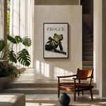 A framed art print featuring a green frog dressed in a black suit with the word 'FROGUE' above it, displayed on a beige wall in a modern, sunlit interior with a wooden chair, plants, and a staircase.