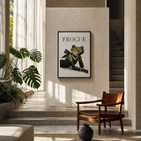 A framed art print featuring a green frog dressed in a black suit with the word 'FROGUE' above it, displayed on a beige wall in a modern, sunlit interior with a wooden chair, plants, and a staircase.