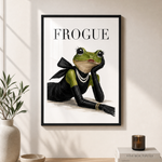 A framed art print featuring a green frog in a black outfit with pearl necklaces and the word 'FROGUE' at the top, displayed on a beige wall above a wooden console table with a vase, bowl, and books.